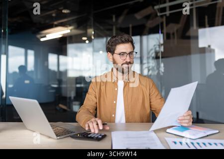 Contabile che lavora diligentemente su un notebook, utilizzando una calcolatrice durante la revisione dei documenti finanziari, la gestione dei budget e l'analisi dei dati in un ambiente di ufficio moderno Foto Stock