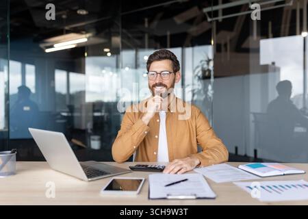 Happy Accountant lavora con documenti finanziari, utilizza laptop e calcolatrice, analizza i grafici delle vendite, siede alla scrivania in un ufficio moderno, tocca il mento e sorride Foto Stock
