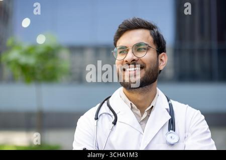 Giovane professionista medico maschile che indossa cappotto bianco e stetoscopio sorridente con sicurezza fuori dalla moderna costruzione ospedaliera, che rappresenta la salute, la professionalità e la competenza Foto Stock