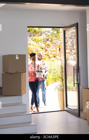 Coppia afroamericana che trasporta piante e scatole che si spostano in una nuova casa, sorridendo con entusiasmo. trasferimento, disimballaggio, felicità, nuovi inizi, lif Foto Stock