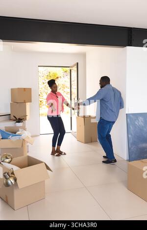 Coppia afroamericana che celebra la nuova casa, ballando tra scatole in movimento in un luminoso salotto. Proprietà, trasferimento, disimballaggio, felicità, gioia, Foto Stock