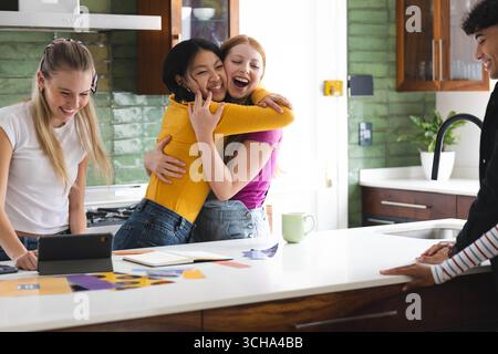 Amici diversi che festeggiano in cucina, si abbracciano e sorridono intorno a tablet e giornali, a casa. Amicizia, celebrazione, tecnologia, giovinezza, togeth Foto Stock
