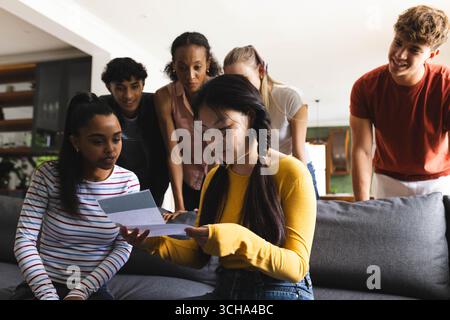 Teen che legge la lettera di accettazione del college sul divano, amici diversi, condividere emozioni, a casa. celebrazione, amicizia, legame, felicità, comunicazione Foto Stock