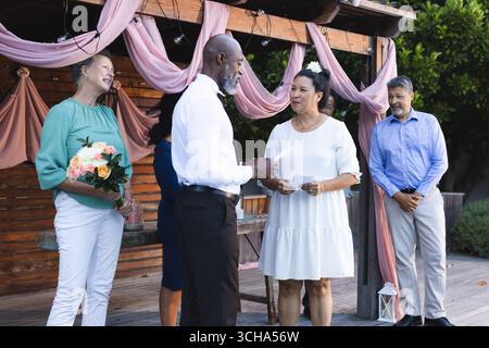 Al matrimonio, scambiandosi i voti in giardino, coppia senior variegata circondata da amici, festeggiando. matrimonio, celebrazione, amore, cerimonia, amicizia, hap Foto Stock