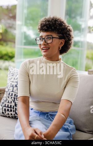 Donna sorridente seduta sul divano di casa, mentre si diverte a conversare in modo rilassato, a casa. Relax, felicità, stile di vita, domestico, tempo libero, informale Foto Stock
