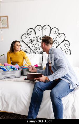 Coppia che prepara la valigia sul letto, si prepara per le vacanze, sorridendo e chiacchierando. Viaggio, bagagli, felicità, preparazione, avventura, eccitazione Foto Stock