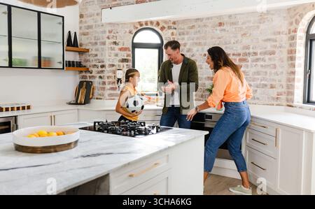 La famiglia gioca con il pallone da calcio in una cucina moderna, trascorrendo momenti di qualità insieme. legame, attività, solidarietà, famiglia, genitorialità, tempo libero Foto Stock