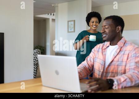 Donna sorridente che tiene il caffè mentre l'uomo lavora con un notebook a casa. Lavoro a distanza, comunicazione, stile di vita, ufficio domestico, relazione, lavoro di squadra Foto Stock