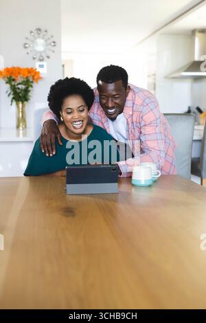 Ridendo coppia afroamericana appoggiata a tavola in una cucina moderna, guardando un tablet con una tazza di caffè. Stile di vita, intimità, felicità, calore, arredamento, mo Foto Stock