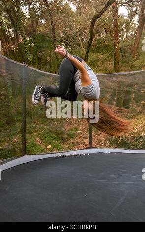 Giovane donna vestita di abbigliamento sportivo che fa un backflip su un trampolino in una foresta all'aperto in montagna, godendosi il momento. Foto Stock