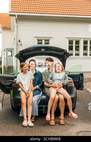 Uomo e donna sorridenti seduti con le figlie nel bagagliaio dell'auto elettrica sul vialetto Foto Stock