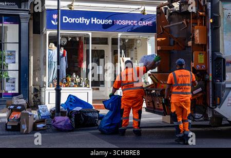 Gli addetti alla raccolta dei rifiuti in uniforme arancione smaltiscono i sacchetti dei rifiuti di fronte a un negozio di beneficenza di ospizio in una giornata di sole a Bath UK Foto Stock