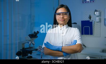 Una scienziata asiatica sicura di sé sta in un laboratorio, le braccia incrociate, indossa l'equipaggiamento protettivo e sorride allo spettatore. Foto Stock