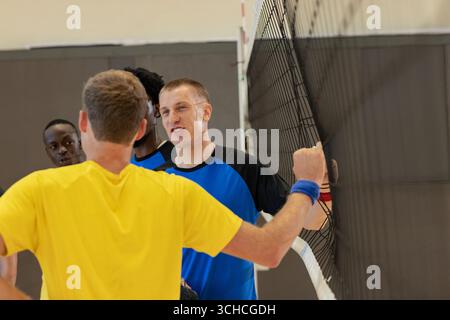 Diversi giocatori di pallavolo maschile che indossano un braccialetto che stringe le mani attraverso la rete in palestra con un palo a strisce. Atleti, lavoro di squadra, celebrazione, competizione, spo Foto Stock