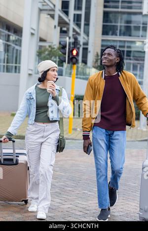 Diversi amici che camminano sulla piazza dell'ufficio tirando le valigie, bevendo un caffè e controllando il telefono. Casual, viaggi, urbano, stile di vita, moderno, avventura, le Foto Stock