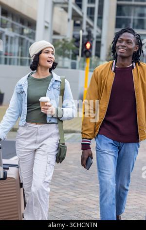 Amici diversi che camminano lungo il marciapiede urbano tenendo in mano valigia, caffè e telefono. Urbano, viaggi, casual, moderno, stile di vita, amicizia, spiegazione Foto Stock