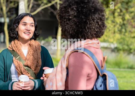 Diverse amiche che chiacchierano tenendo tazze da caffè con la borsa a tracolla e lo zaino nel campus. Casual, Friendship, urban, lifestyle, vivace, motore Foto Stock