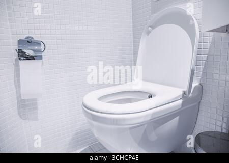 La ciotola della toilette bianca pulita con coperchio aperto in un bagno moderno con piccole piastrelle bianche e un rotolo di carta igienica sul muro Foto Stock