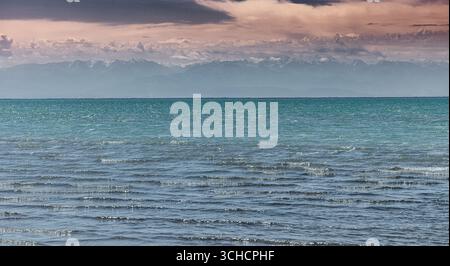 L'acqua calmo che lambisce dolcemente la riva, con le montagne Tian Shan visibili in lontananza sotto un cielo nuvoloso Foto Stock