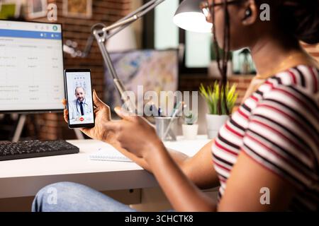 Donna nera che tiene il cellulare e ha parlato con un medico professionista in videochiamata. Individuo afroamericano femminile che condivide gli aggiornamenti sulla salute con il medico caucasico durante l'incontro virtuale. Foto Stock