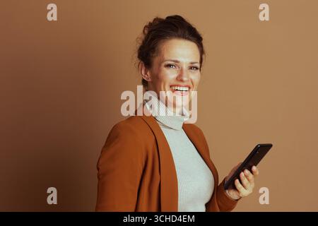 Una donna d'affari sicura e sorridente, con blazer e dolcevita, tiene lo smartphone, guardando direttamente lo spettatore. Questo ritratto cattura la professione moderna Foto Stock