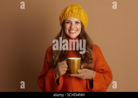 Una donna sorridente con berretto giallo e maglione arancione tiene la tazza gialla con entrambe le mani, guardando direttamente la fotocamera. L'atmosfera calda e allegra suggerisce comfort Foto Stock