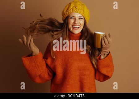 Una donna allegra con i capelli marroni che scorrono, con un cappello in maglia gialla e un maglione arancione, ride gioiosamente mentre tiene in mano una tazza gialla di senape. Il suo vibran Foto Stock