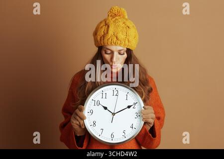 Una donna con un berretto giallo e un maglione arancione con bucchi divertenti, tenendo un grande orologio da parete su un caldo sfondo marrone. I suoi occhi chiusi suggeriscono formiche Foto Stock