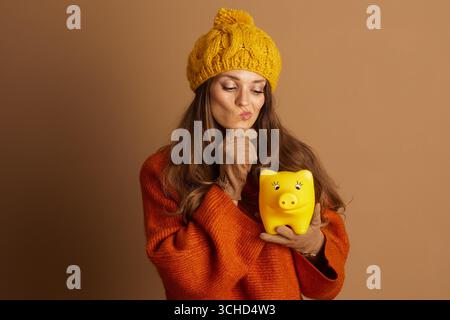 Una donna contemplativa in maglione arancione e berretto giallo posa con salvadanaio, mano sul mento e labbra borsate. Esamina attentamente i suoi risparmi, Foto Stock