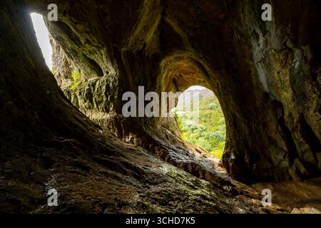 La luce del sole che entra attraverso le aperture in una grande grotta illumina l'interno roccioso e rivela una vista su una lussureggiante valle Foto Stock