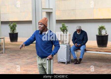 Viaggiatori afroamericani che tirano le valigie dall'altra parte della piazza mentre uno controllava il telefono sulla panchina Foto Stock
