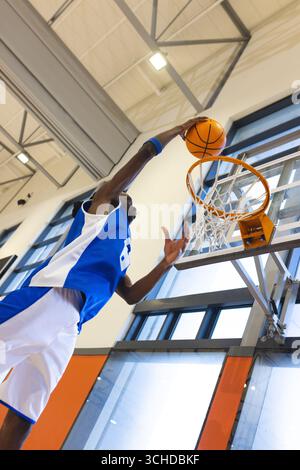 Uomo afroamericano che salta in Jersey blu che fa schifo da basket e indossa una palestra con backboard in vetro Foto Stock