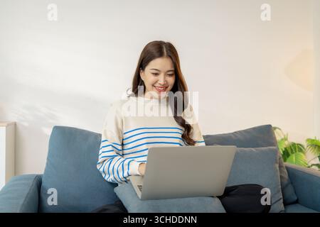 Una giovane donna è seduta comodamente su un divano utilizzando un computer portatile, sorridendo con concentrazione e gioia. Sembra impegnata nel suo lavoro o nella comunicazione online, crea Foto Stock