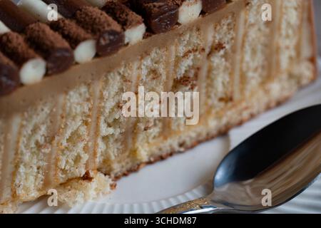 Primo piano di una fetta di pan di Spagna a strati con ripieno cremoso, ricoperta da decorazioni di cioccolato e cacao spolverato in polvere, servito con un cucchiaio Foto Stock