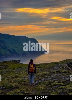 Un escursionista si erge su una scogliera erbosa, guardando il tramonto mozzafiato sulle tranquille acque delle Isole Faroe. Il suggestivo paesaggio presenta montagne ripide e cieli vibranti. Foto Stock