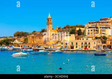 Vista colorata di Marsaskala, Malta, con barche che galleggiano sul mare blu brillante e storici edifici in pietra lungo il lungomare. Un giro panoramico sulla costa Foto Stock
