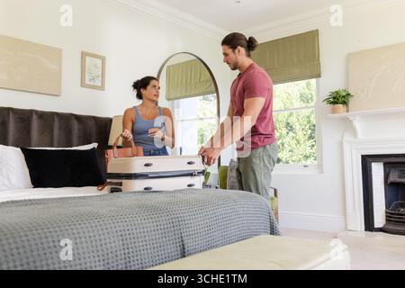 Disimballare la coppia organizzando i vestiti sul letto in una camera da letto luminosa arredata, con valigia e borsa Foto Stock