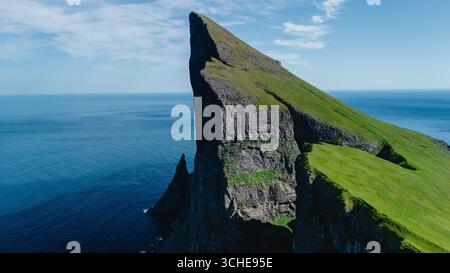 Una spettacolare scogliera sorge bruscamente dalle vibranti acque blu dell'Oceano Atlantico nelle Isole Faroe. La lussureggiante vegetazione contrasta con i bordi rocciosi, creando un panorama naturale mozzafiato. Foto Stock