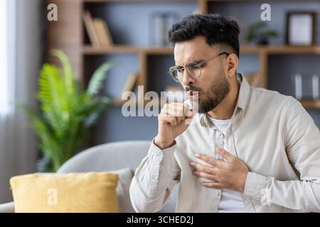 L'uomo seduto sul divano sembra tossire e tenere il petto in mano. Indossando occhiali e abbigliamento casual, sembra preoccupato per la salute. Ambiente interno con Foto Stock