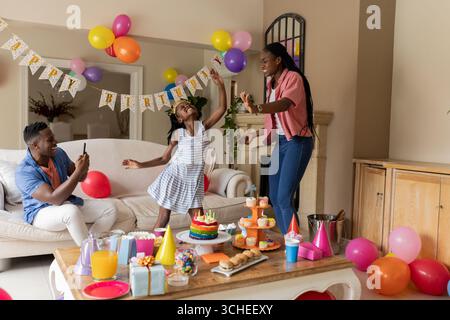 Famiglia che festeggia il compleanno a casa, figlia che balla allegramente con i genitori che esultano Foto Stock
