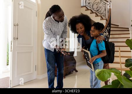 Genitori afroamericani che aiutano il figlio con zaino e tablet all'ingresso di casa Foto Stock