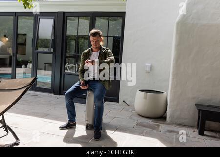 Uomo seduto in valigia a controllare il tempo su uno smartwatch nel patio soleggiato Foto Stock