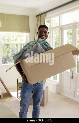 Uomo afro-americano che trasporta una scatola di cartone in un luminoso e arioso soggiorno, con cuscini e coperte Foto Stock