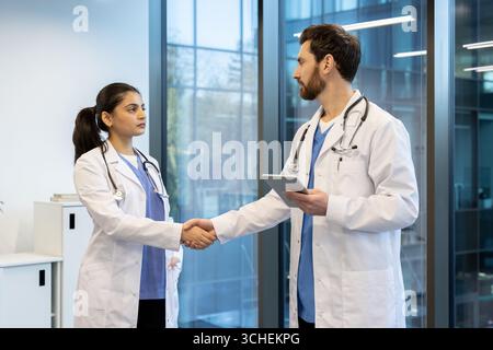 Due medici, un uomo e una donna, stringono la mano in un moderno corridoio ospedaliero, dimostrando collaborazione medica e lavoro di squadra, con il medico maschile che tiene in mano una tavoletta digitale Foto Stock
