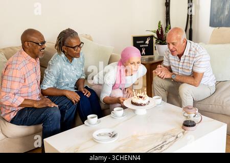 Amici anziani che festeggiano il compleanno a casa, donna che fa saltare le candele sulla torta Foto Stock