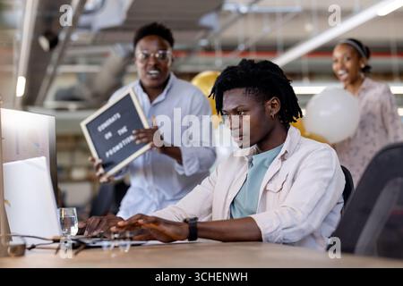 Un giovane che scrive sul laptop in ufficio mentre colleghi diversi festeggiano nelle vicinanze Foto Stock