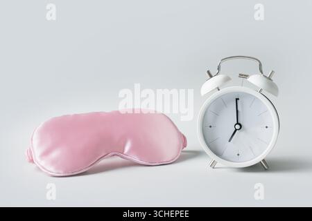 Maschera dormiente rosa e sveglia bianca su sfondo grigio. Concetto di sonno, insonnia, assistenza sanitaria, routine quotidiana e ritmi circadiani. Foto Stock