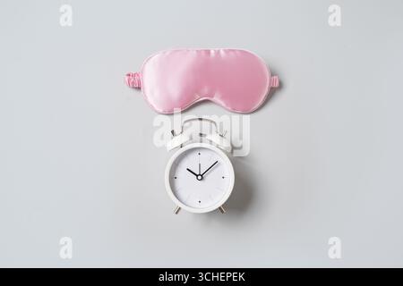 Maschera dormiente rosa e sveglia bianca su sfondo grigio. Concetto di sonno, insonnia, assistenza sanitaria, routine quotidiana e ritmi circadiani. Copia spazio. Foto Stock