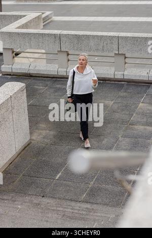 Donna anziana che cammina attraverso la piazza piastrellata da ringhiere di metallo che tengono una tazza di caffè e una borsa verde Foto Stock