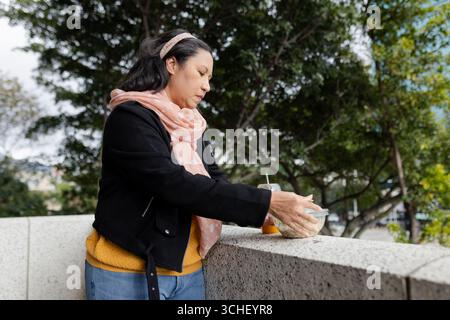 Donna asiatica matura che indossa sciarpa rosa collocando insalata ciotola, bevanda su ringhiera della terrazza, copia spazio Foto Stock